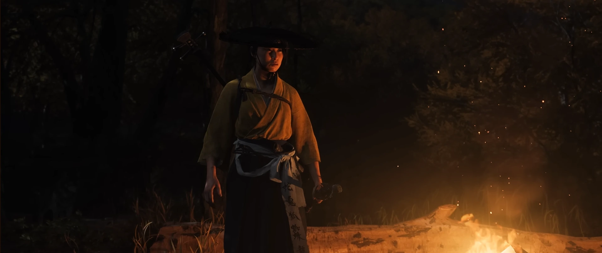 A character in traditional Japanese attire and a wide-brimmed hat stands by a campfire in a dark forest scene from Ghost of Yōtei.
