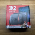 The retail box for the Nintendo Switch 2, prominently displaying the console, detachable Joy-Con controllers in a new design, and advertising a full game download for 'Mario Kart World'. The box is primarily red and black, with white text, and rests on a wooden surface.