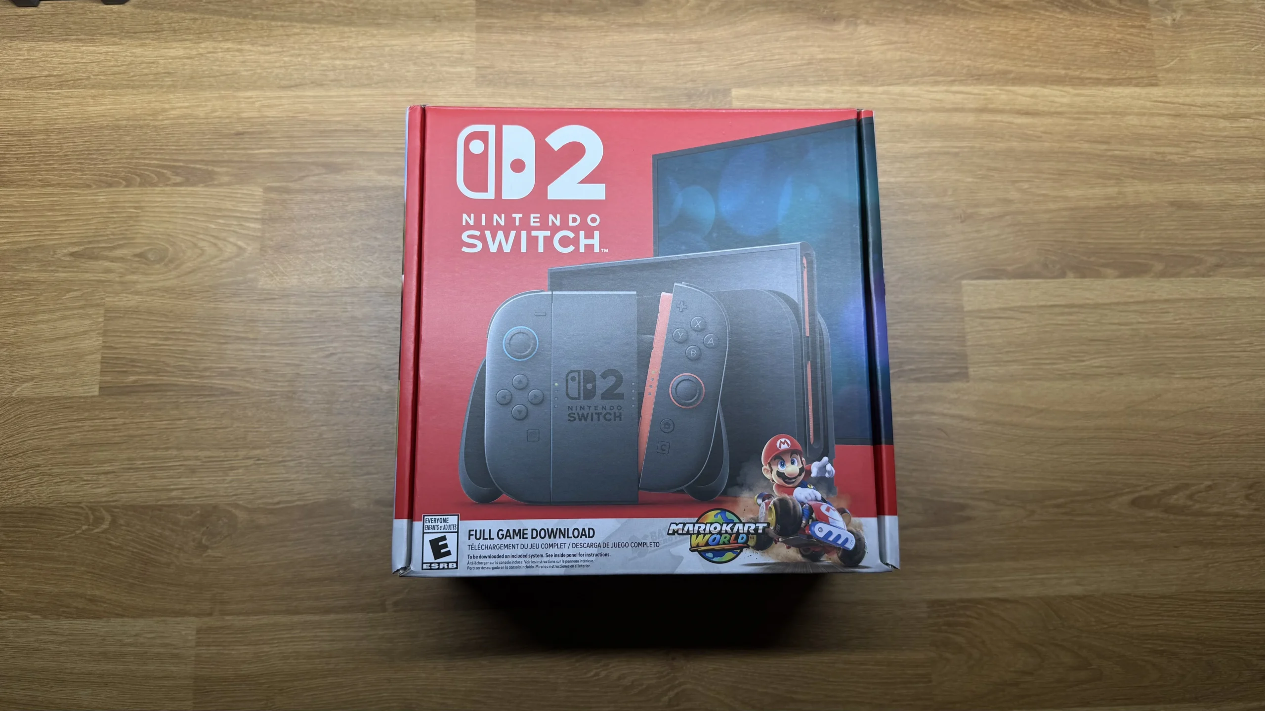 The retail box for the Nintendo Switch 2, prominently displaying the console, detachable Joy-Con controllers in a new design, and advertising a full game download for 'Mario Kart World'. The box is primarily red and black, with white text, and rests on a wooden surface.