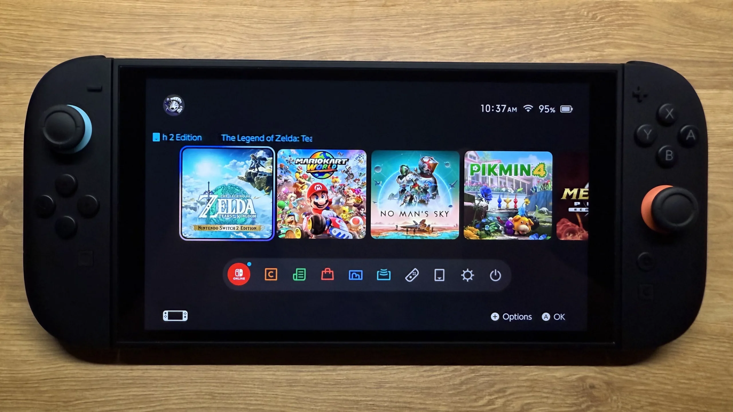 A photo of the Nintendo Switch 2 console, showing its home screen interface. The screen displays a grid of game icons, including 'The Legend of Zelda: Tears of the Kingdom', 'Mario Kart 8 Deluxe', 'No Man's Sky', and 'Pikmin 4', along with system icons for the eShop, Album, Controllers, and System Settings at the bottom. The top right shows the time, battery life, and Wi-Fi signal.
