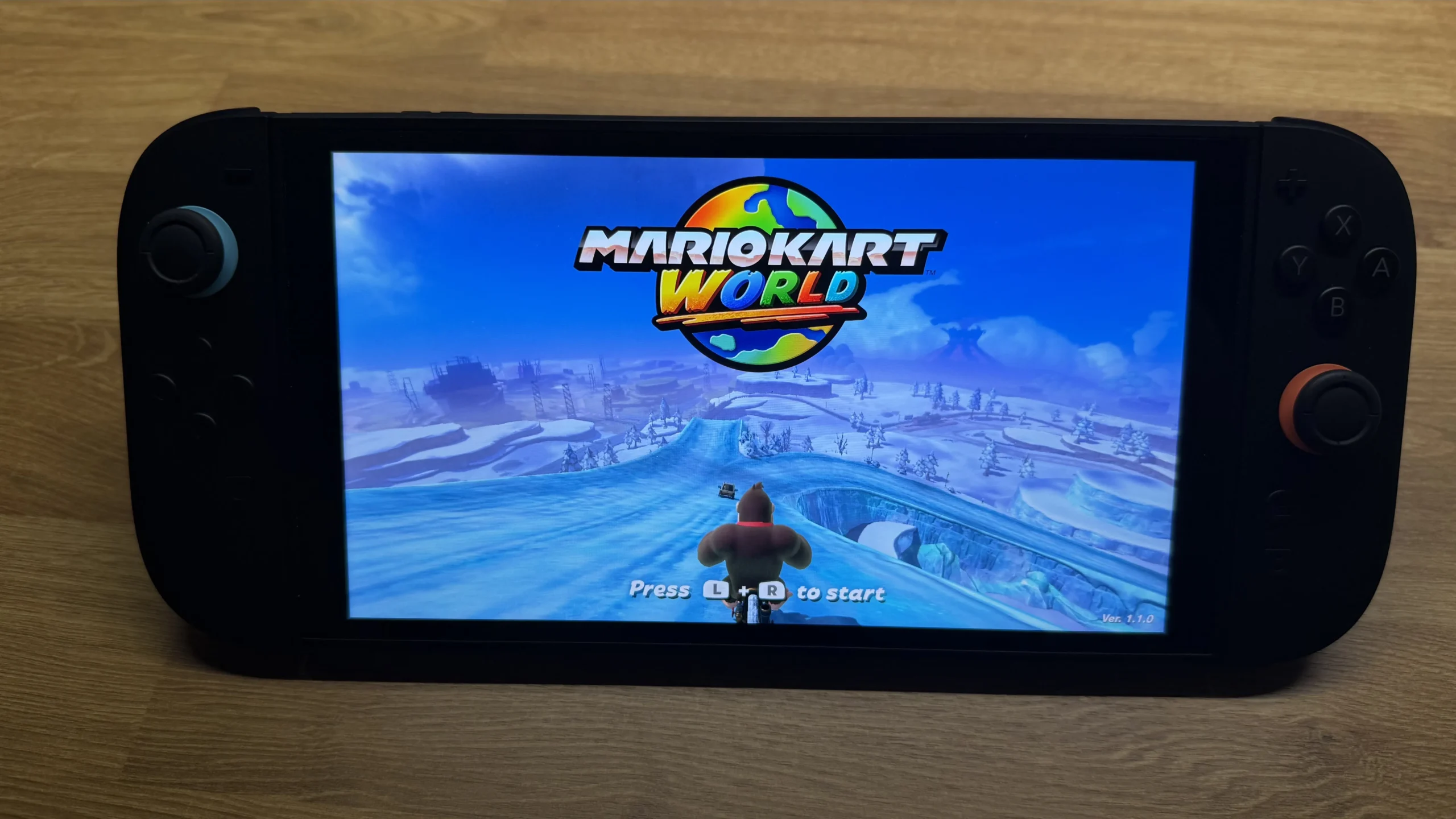 A close-up view of the Nintendo Switch 2 handheld console, powered on and showing the "Mario Kart World" title screen. The game features a character on a snowy track under a blue sky, with the text "Press L + R to start" visible. The console itself is black with blue and orange accents on the Joy-Cons.