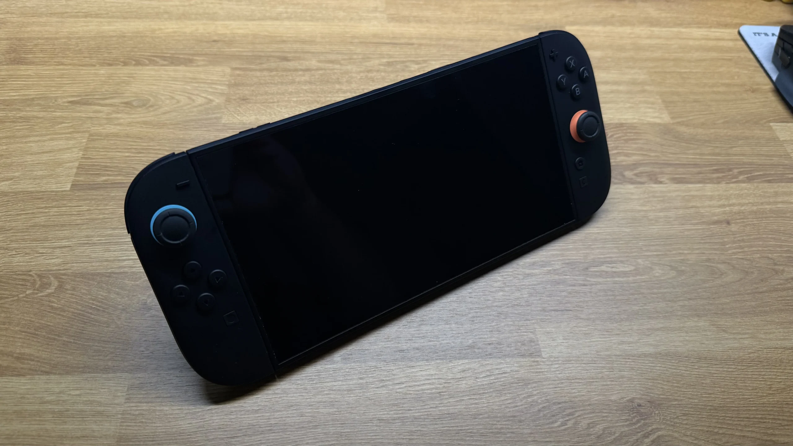 A close-up photo of the Nintendo Switch 2 console with black Joy-Cons, featuring a prominent, glossy black screen and orange joystick accents.