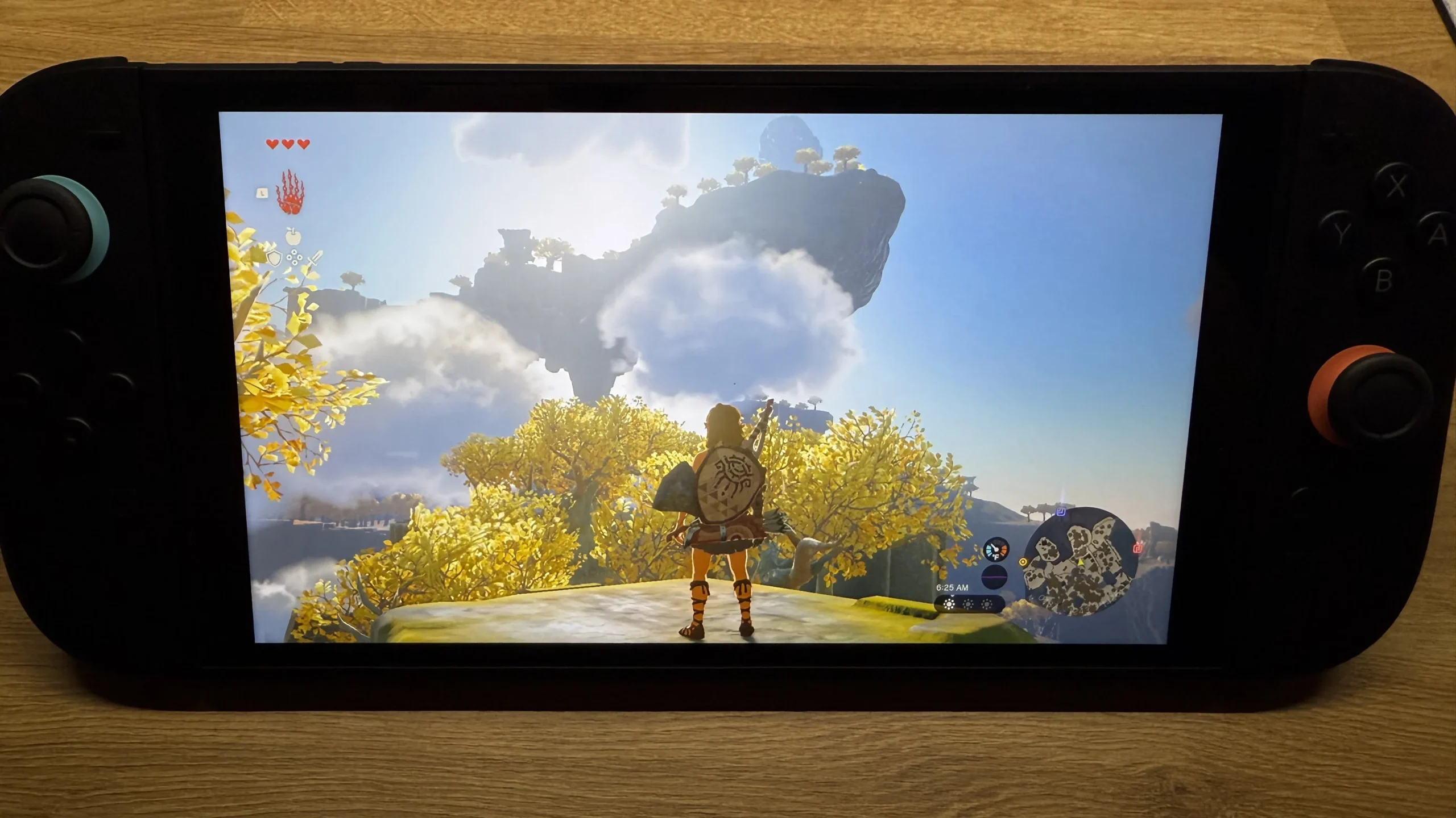 A close-up of the Nintendo Switch 2, held horizontally, showing a vibrant scene from The Legend of Zelda: Tears of the Kingdom on its screen. The character Link stands on a cliff overlooking a fantastical landscape with a floating island in the sky and golden trees, highlighting the game's graphics on the new console. The Joy-Cons are black with blue and orange accents.