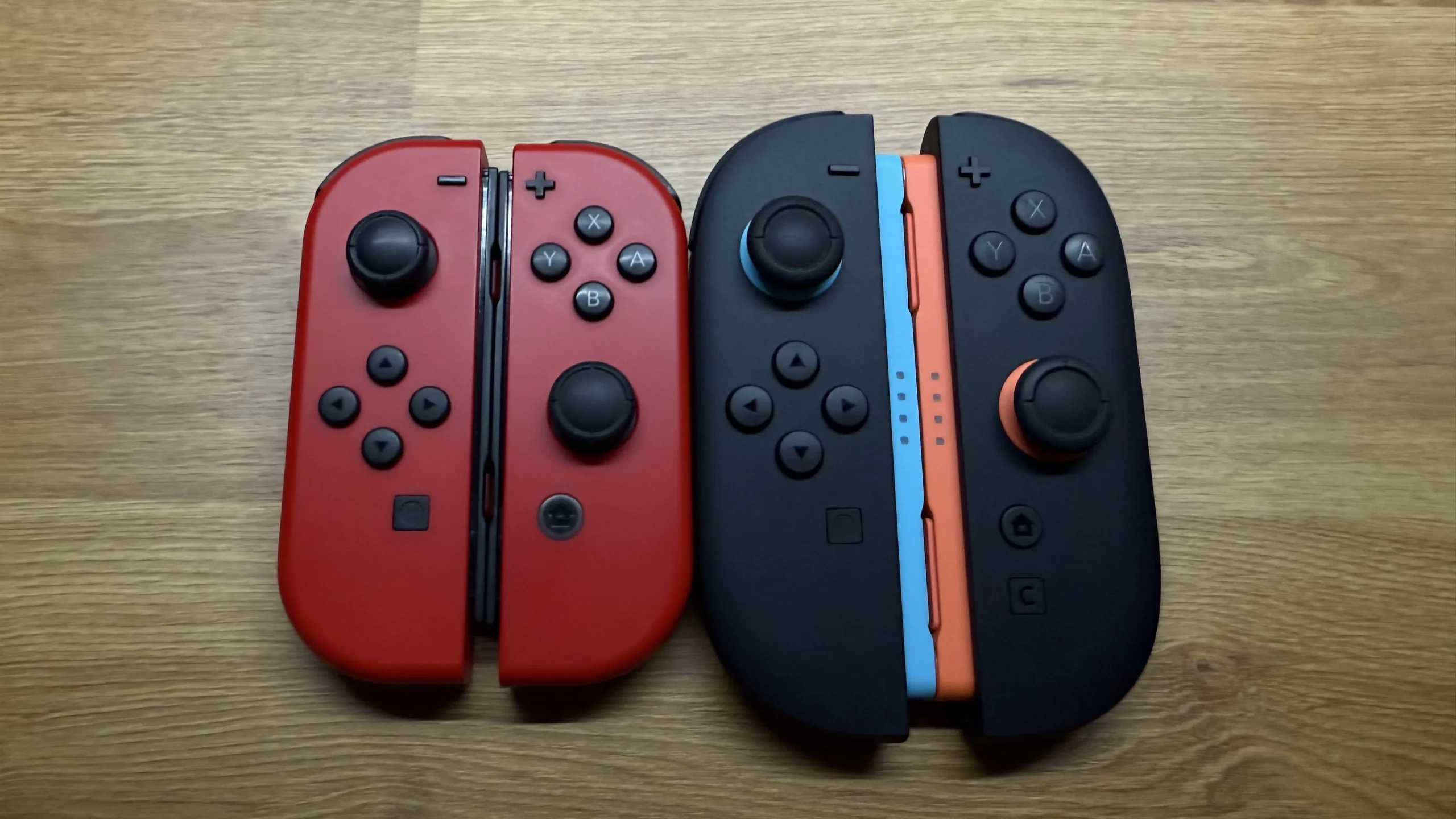 A close-up overhead shot comparing two sets of Joy-Cons. On the left are a pair of red Joy-Cons from the original Nintendo Switch, and on the right are a pair of black Joy-Cons with orange and blue inner rails the Nintendo Switch 2, highlighting a potential size and design change between the two.